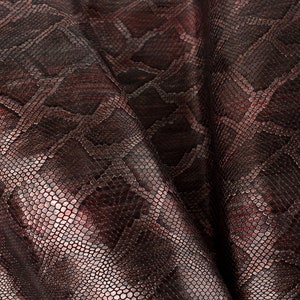 Könnte beinhalten: Nahaufnahme eines dunkelbraunen und burgunderfarbenen Schlangenlederstoffs mit einem metallischen Schimmer. Der Stoff hat ein strukturiertes, schuppiges Muster.