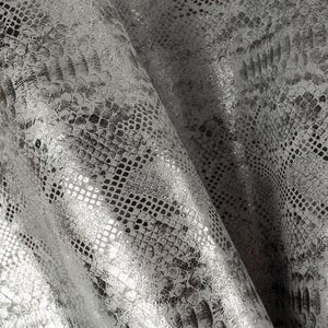Puede incluir: Tela de piel de serpiente plateada con un patrón texturizado y estampado. La tela tiene un brillo metálico y un ligero brillo.