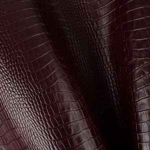 May include: Close-up of a burgundy faux crocodile leather fabric with a textured, embossed pattern.