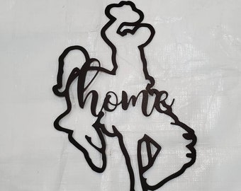 May include: Black metal wall art featuring a silhouette of a cowboy riding a bucking bronco. The word "home" is written in cursive across the center of the design. This decorative piece is suitable for indoor or outdoor use.