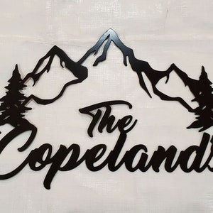 May include: Black metal wall art featuring a mountain range, pine trees, and the family name "The Copelands" in a cursive font. The artwork is designed to be mounted on a wall and adds a rustic touch to home decor.