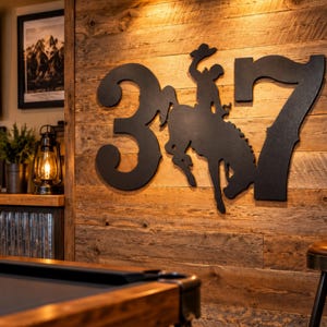 Könnte beinhalten: Ein großes, mattschwarzes Wandobjekt aus Metall, das die Zahl 3, die Silhouette eines Reiters auf einem bockenden Pferd und die Zahl 7 zeigt. Das Schild ist an einer Wand aus horizontalen, verwitterten braunen Holzplanken befestigt.