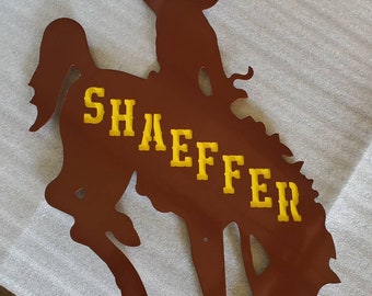 May include: Brown metal cutout of a cowboy riding a bucking bronco. The word "Shaeffer" is in yellow letters across the horse's body. The cowboy is wearing a hat and has one arm raised.