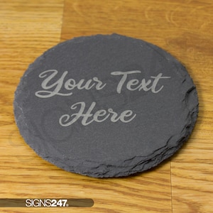 May include: A round, dark grey slate coaster with a rough, natural edge. The surface is engraved with the text "Your Text Here" in a cursive script font. The coaster has a flat, circular shape and a textured, matte finish across its surface.
