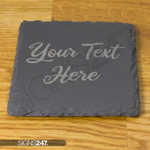 May include: A square dark grey slate coaster with rough, uneven edges. The surface features the engraved text "Your Text Here" in a cursive script font. The coaster has a natural stone texture and a matte finish, providing a sturdy base.