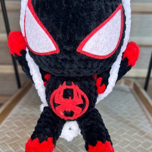 May include: A black and red crocheted plush toy of Spider-Man swinging on a white string. The toy has a red spider symbol on its chest and white eyes with red outlines.