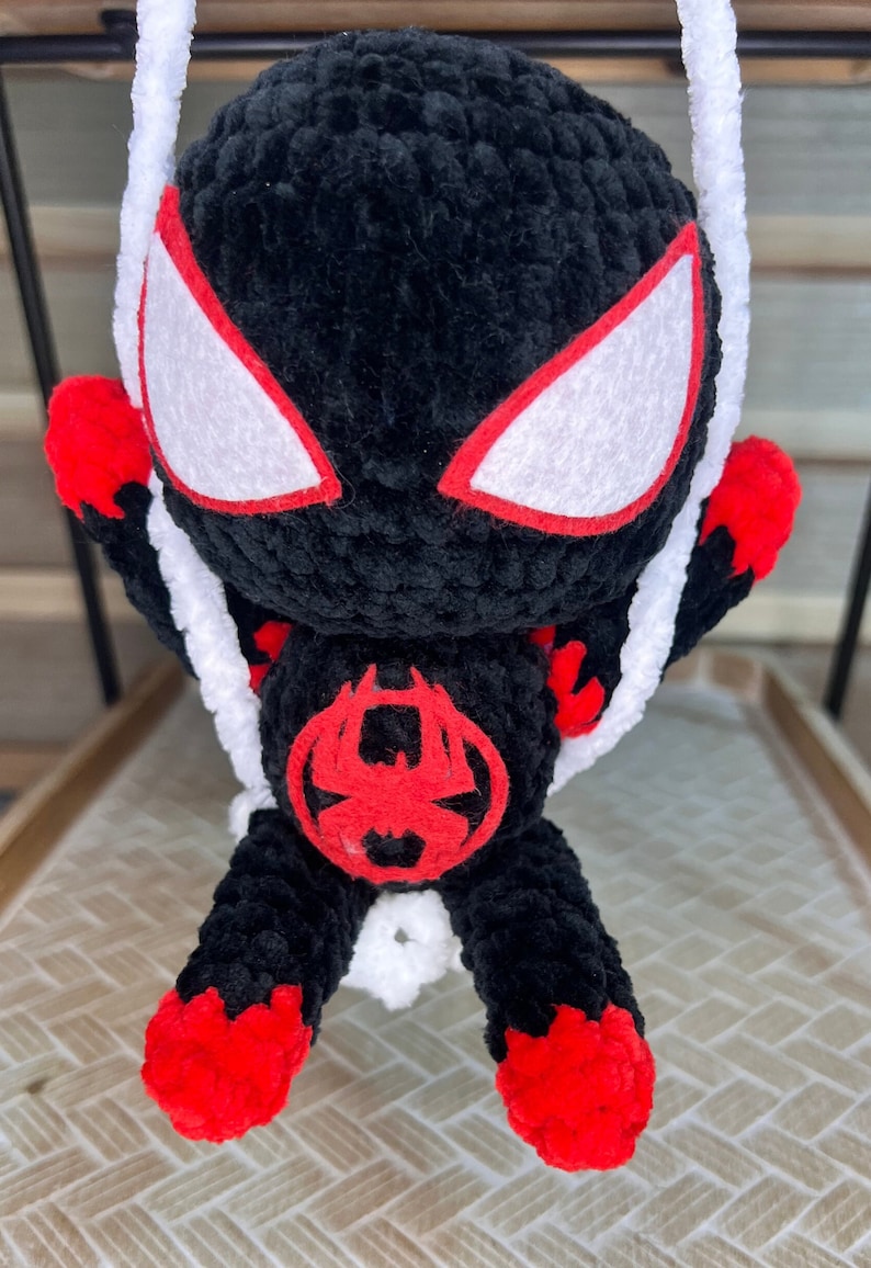 May include: A black and red crocheted plush toy of Spider-Man swinging on a white string. The toy has a red spider symbol on its chest and white eyes with red outlines.