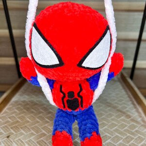 May include: A red and blue crocheted Spiderman toy hanging from a white string. The toy has a black spider symbol on its chest and white eyes.