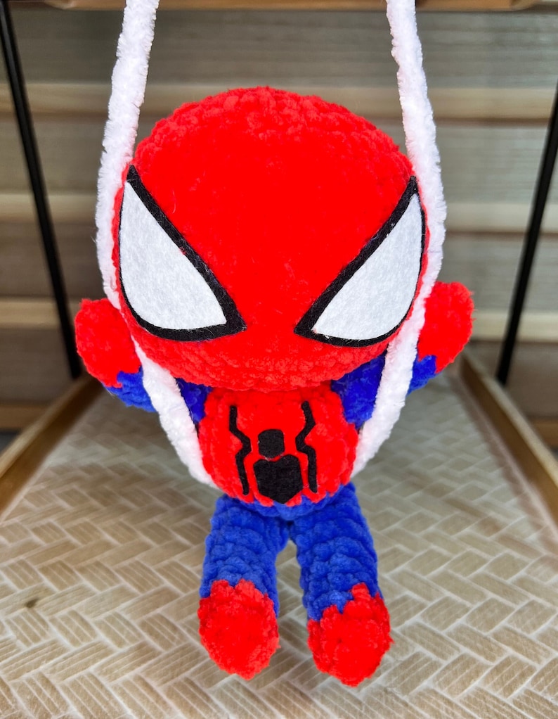 May include: A red and blue crocheted Spiderman toy hanging from a white string. The toy has a black spider symbol on its chest and white eyes.