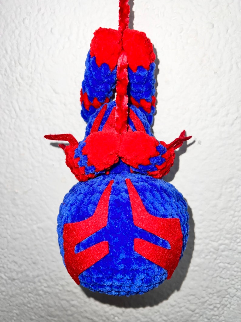 May include: A blue and red crocheted plush toy shaped like a spider hanging from a white ceiling. The spider has a red and blue pattern on its body.
