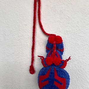 May include: A blue and red crocheted toy with a long red cord. The toy has a round body with red accents and a long, thin tail.