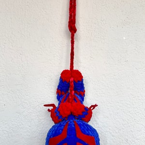 May include: A blue and red crocheted plush toy hanging from a red string. The toy is shaped like a spider with a round body and eight legs.