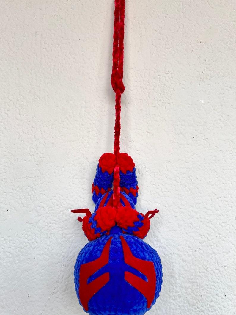 May include: A blue and red crocheted plush toy hanging from a red string. The toy is shaped like a spider with a round body and eight legs.