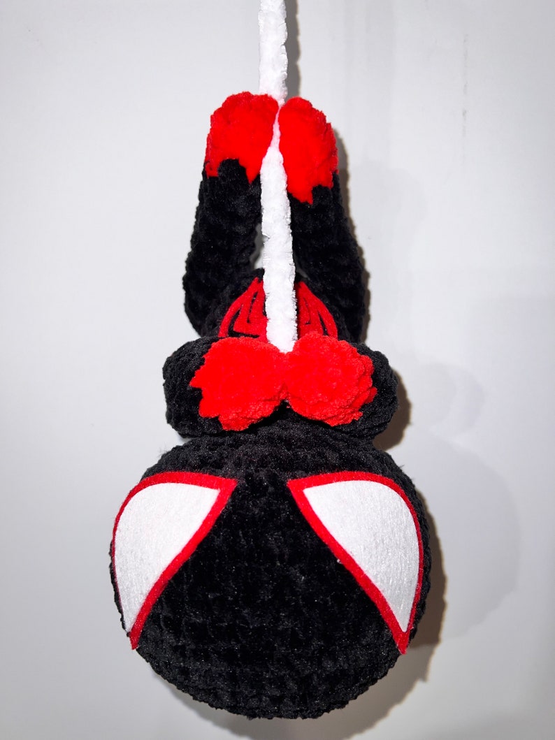 May include: Black and red crocheted plush toy hanging from a white string. The toy has a spider-like design with white eyes.