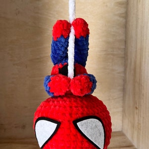 May include: A red and blue crocheted Spiderman toy hanging from a white string. The toy has white felt eyes and a red and blue crocheted body.
