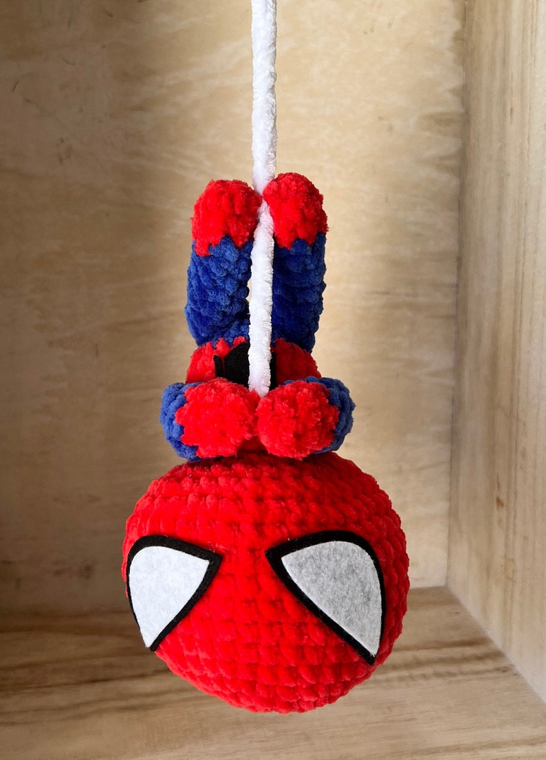 May include: A red and blue crocheted Spiderman toy hanging from a white string. The toy has white felt eyes and a red and blue crocheted body.