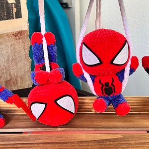 May include: Four crocheted Spider-Man figures hanging from white strings. The figures are red, blue, and black with white eyes. The figures are hanging from a wooden surface.
