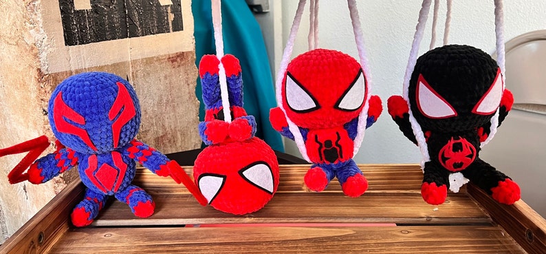 May include: Four crocheted Spider-Man figures hanging from white strings. The figures are red, blue, and black with white eyes. The figures are hanging from a wooden surface.
