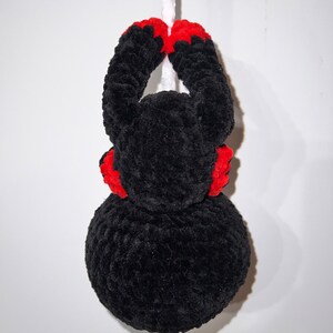 May include: Black crocheted spider hanging from a white string. The spider has red accents on its legs and head.