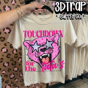 Op de afbeelding: Een crèmekleurig T-shirt met korte mouwen en een afbeelding van een roze panterkop met stervormige ogen. De tekst TOUCHDOWN for the Tata's staat in felroze letters. Het ontwerp is omlijnd door een zwart rechthoekig kader.