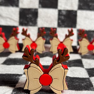 以下が含まれることがあります： クリスマスをテーマにしたリボン装飾のコレクション。各リボンは赤い中心部、ベージュのリボン、茶色のトナカイの角が特徴です。リボンは白と黒のチェック柄の表面に配置されています。