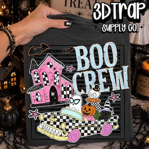 Op de afbeelding: Een zwart t-shirt met een grafische print van een roze spookhuis en een zwart-wit geblokte auto met een geest, een pompoen en een kat. De tekst BOO CREW is in lichtblauw gedrukt en het logo 3DTRAP SUPPLY CO. staat bovenaan.
