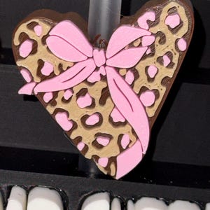 May include: A heart-shaped charm with a brown border, tan leopard print, and pink bow. The charm is a decorative accessory. The word "BAD" is visible at the bottom of the image.