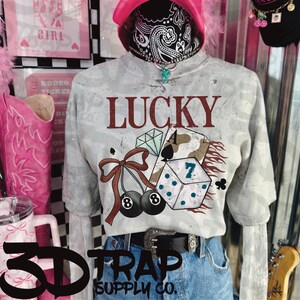 Op de afbeelding: Een grijs tie-dye sweatshirt met het woord "LUCKY" in kastanjebruine letters. De afbeelding bevat dobbelstenen, speelkaarten, kersen en het nummer 7. Het shirt wordt gecombineerd met een spijkerbroek en een zwarte riem met een zilveren gesp. De tekst "3D TRAP SUPPLY CO." staat onderaan.