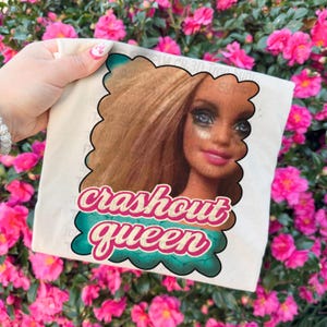 May include: A beige t-shirt featuring a doll's face with the text "crashout queen" in a teal and pink design. The doll has blonde hair, blue eyes, and pink lipstick. The t-shirt is held up against a backdrop of pink flowers.
