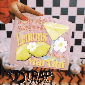 May include: A pink cocktail napkin with a lemon and martini design. The napkin features the text "When life gives you lemons, make a Martini." The design includes yellow lemons, a martini glass, and white flowers.