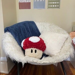 Op de afbeelding: Een witte nep bont stoel met een rood en wit gebreid paddenstoel kussen en een wit kussen. De stoel staat op een houten vloer.