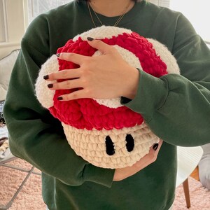 Op de afbeelding: Een gehaakt kussen in de vorm van een paddenstoel met een rood-witte bovenkant en een cr&egrave;mekleurige onderkant. Het kussen heeft twee zwarte ogen.