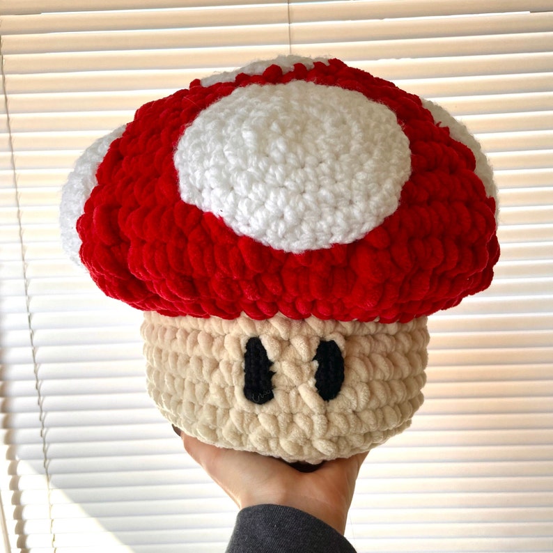 Op de afbeelding: Een gehaakt pluche paddenstoel speelgoed met een rood-witte dop en een beige lichaam met zwarte ogen.