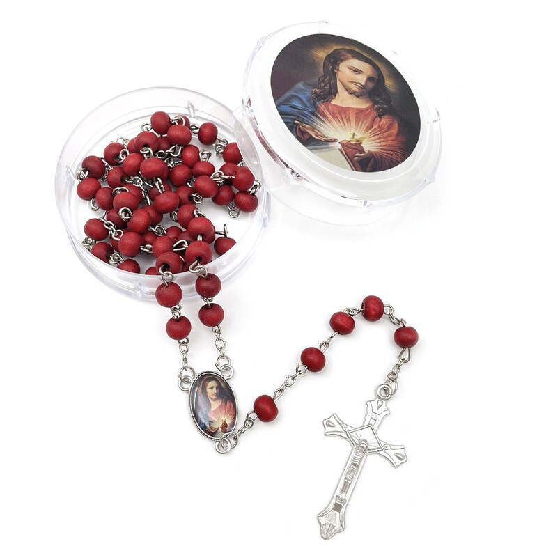 Puede incluir: Un rosario de cuentas de madera roja con un crucifijo y una cadena de conexi&oacute;n plateados. El rosario se muestra en una caja de pl&aacute;stico transparente con una imagen de Jes&uacute;s. Las cuentas son de color rojo oscuro.