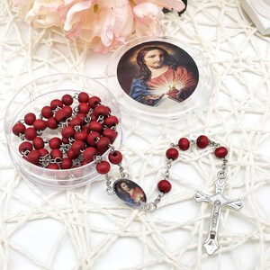 Puede incluir: Un rosario con cuentas de madera rojas y un crucifijo plateado. El rosario se muestra con un recipiente de pl&aacute;stico transparente que contiene m&aacute;s cuentas y una peque&ntilde;a imagen de Jes&uacute;s. El fondo es una superficie blanca texturizada.