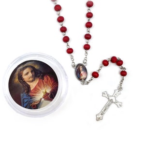 Puede incluir: Un rosario de cuentas de madera roja con un crucifijo plateado y una cadena de conexi&oacute;n. Hay un colgante ovalado con una imagen religiosa. Un recipiente redondo transparente contiene una imagen religiosa.