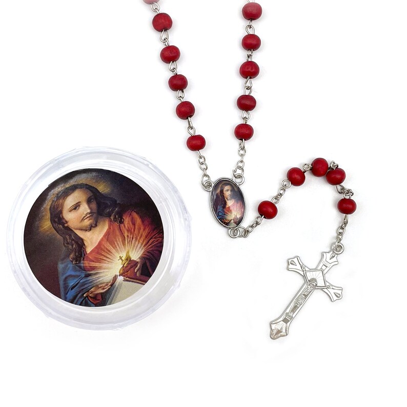 Puede incluir: Un rosario de cuentas de madera roja con un crucifijo plateado y una cadena de conexi&oacute;n. Hay un colgante ovalado con una imagen religiosa. Un recipiente redondo transparente contiene una imagen religiosa.