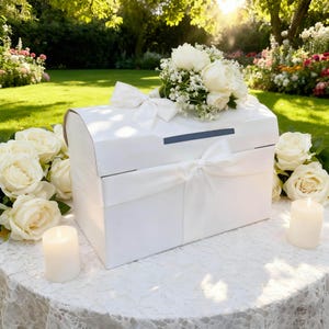 May include: A white, rectangular card box with a curved top, adorned with white ribbon and a bouquet of white roses and baby's breath. The box sits on a lace-covered table, surrounded by white roses and lit candles, with a garden backdrop.