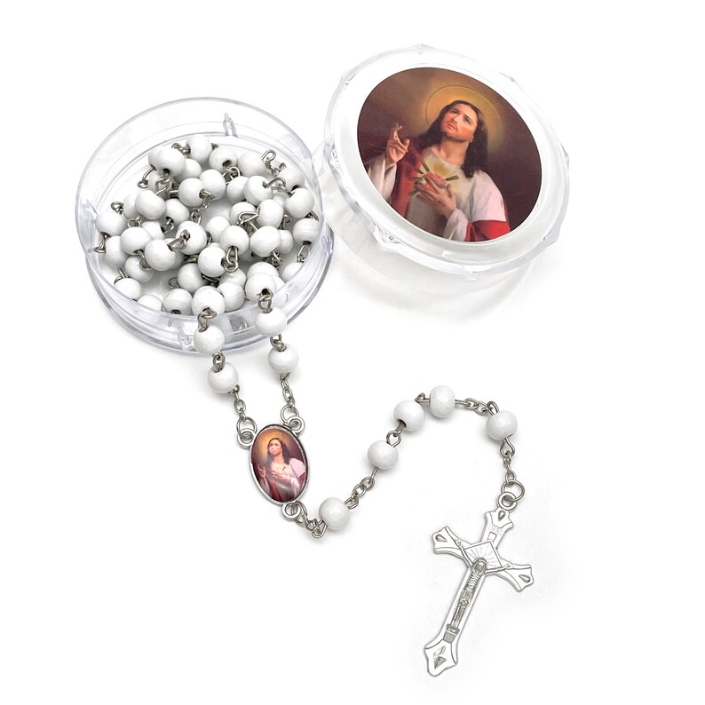 Puede incluir: Un rosario blanco con una cadena y un crucifijo plateados. Las cuentas del rosario son redondas y se guardan en un recipiente de pl&aacute;stico transparente. La tapa del recipiente presenta una imagen de Jes&uacute;s. Tambi&eacute;n hay un colgante ovalado con la imagen de una figura religiosa.