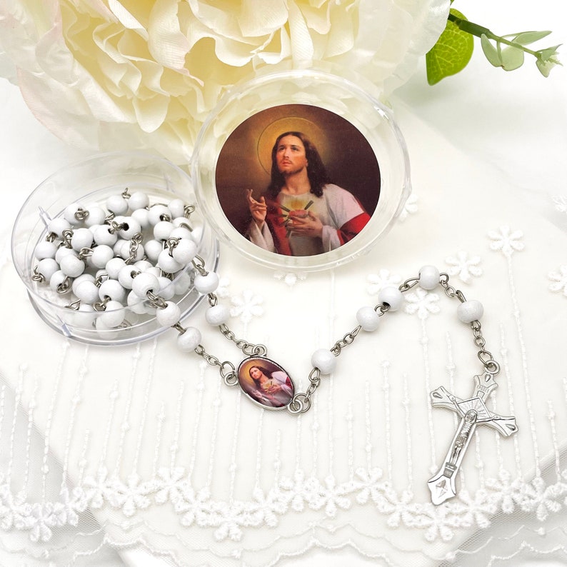 Puede incluir: Rosario de cuentas blancas con crucifijo plateado y pieza central ovalada. El rosario se muestra con un recipiente de pl&aacute;stico transparente que contiene cuentas adicionales y una imagen de Jes&uacute;s. Fondo de encaje blanco.