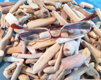 May include: A pair of reading glasses with dark red frames and clear lenses resting on a large pile of smooth, light-coloured driftwood pieces. The glasses feature a rectangular shape and are positioned centrally atop the scattered wooden fragments.