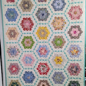 May include: A handmade quilt with a repeating floral pattern. The quilt features numerous colorful floral medallions set against a white background with a light blue grid pattern. The edges are finished with a light blue border.