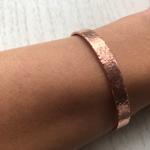 May include: A hammered copper cuff bracelet. The bracelet has a textured surface and a warm, reddish-brown colour. The cuff is worn on the wrist and has a simple, elegant design. The bracelet is a piece of jewellery.