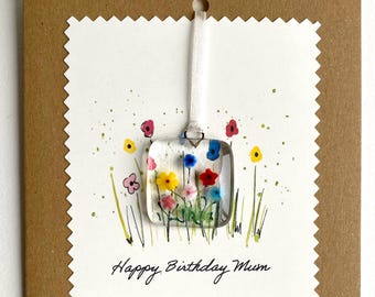 May include: A white card with a scalloped edge and a white ribbon. The card has a hand-drawn image of a field of flowers with a small glass square charm with colourful flowers inside. The text "Happy Birthday Mum" is written on the card.
