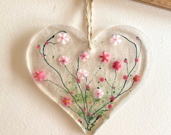 May include: A clear glass heart-shaped ornament with a pink and green floral design. The ornament is hanging from a string.