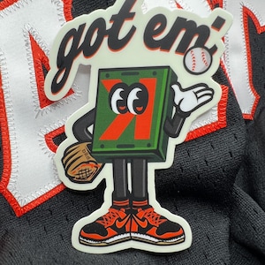 May include: A sticker featuring a cartoon box character with a baseball glove and a baseball. The box is green with an orange "K" and the text "got em'" in a script font. The character is wearing orange and black sneakers.