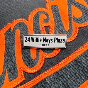 May include: A white rectangular sign with black text that reads "24 Willie Mays Plaza (KING)".