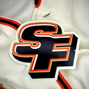 May include: A die-cut sticker featuring the letters "SF" in a bold, stylized design. The letters are black with orange and gold accents, outlined in white. The sticker is placed on a white polo shirt with orange and black trim.