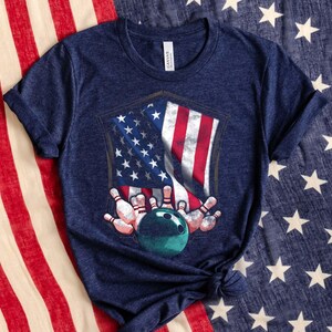 May include: Navy blue t-shirt featuring a graphic of the American flag, bowling pins, and a bowling ball. The design is in a shield shape. The shirt is made of a soft material.