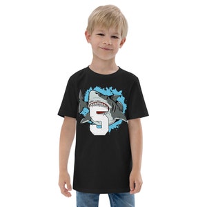 May include: Black short-sleeved t-shirt with a graphic of a shark biting a number 5. The shark is white and gray with a blue background.
